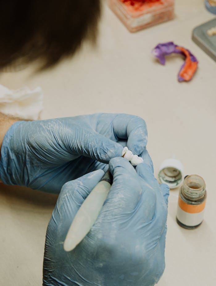 Crop anonymous orthodontist in latex gloves creating fake teeth using dental tools while sitting at table in dental clinic